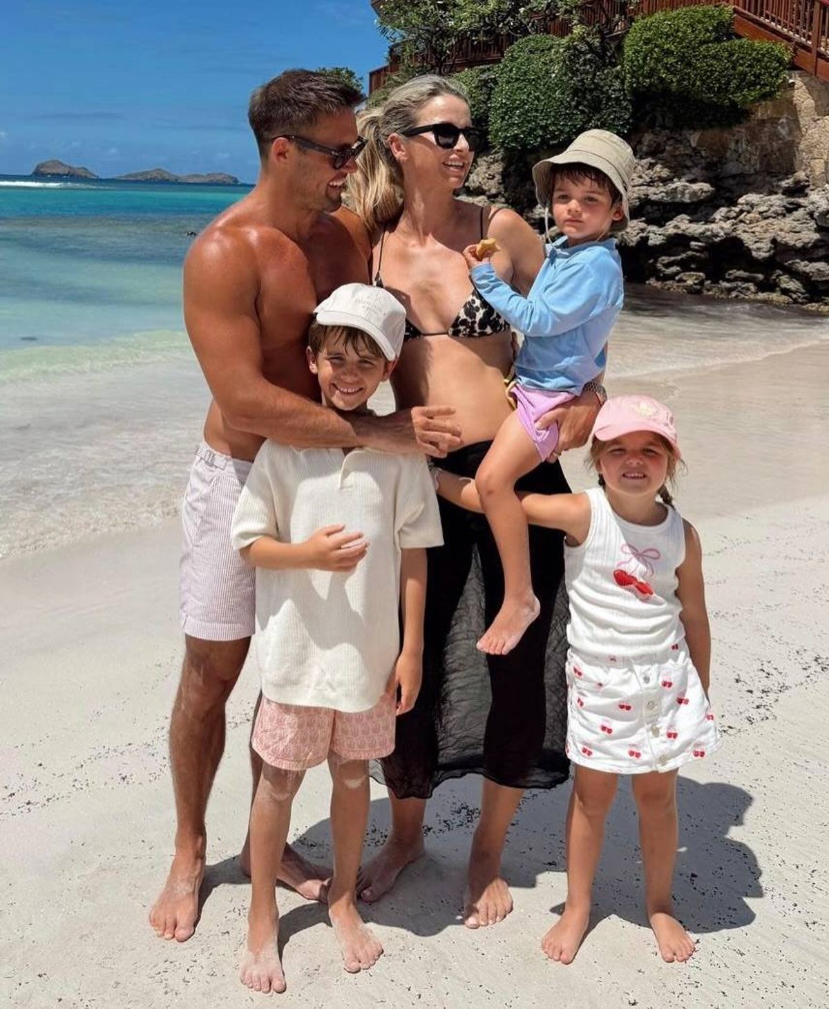 A family of five, consisting of two adults and three children, is standing close together on a sandy beach. They are all wearing hats and seem to be posing for a photograph. The man is shirtless, while the woman is in a bikini. The children are wearing casual beach attire. The beach has a clear blue ocean and some rocks visible in the background.