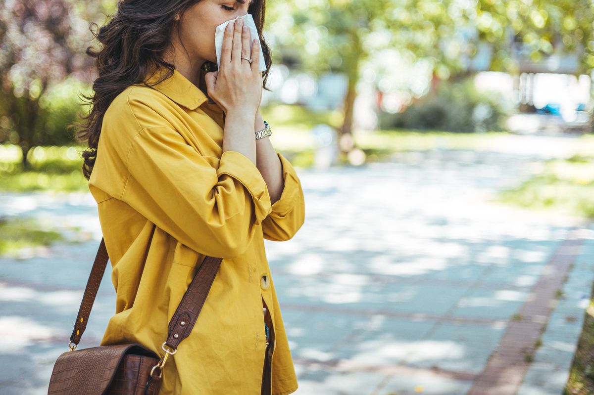 Close-up of a young woman blowing nose with tissue paper at the park. Woman with with allergy symptom blowing nose. Young pretty woman sneezing in front of blooming tree. Spring allergy concept