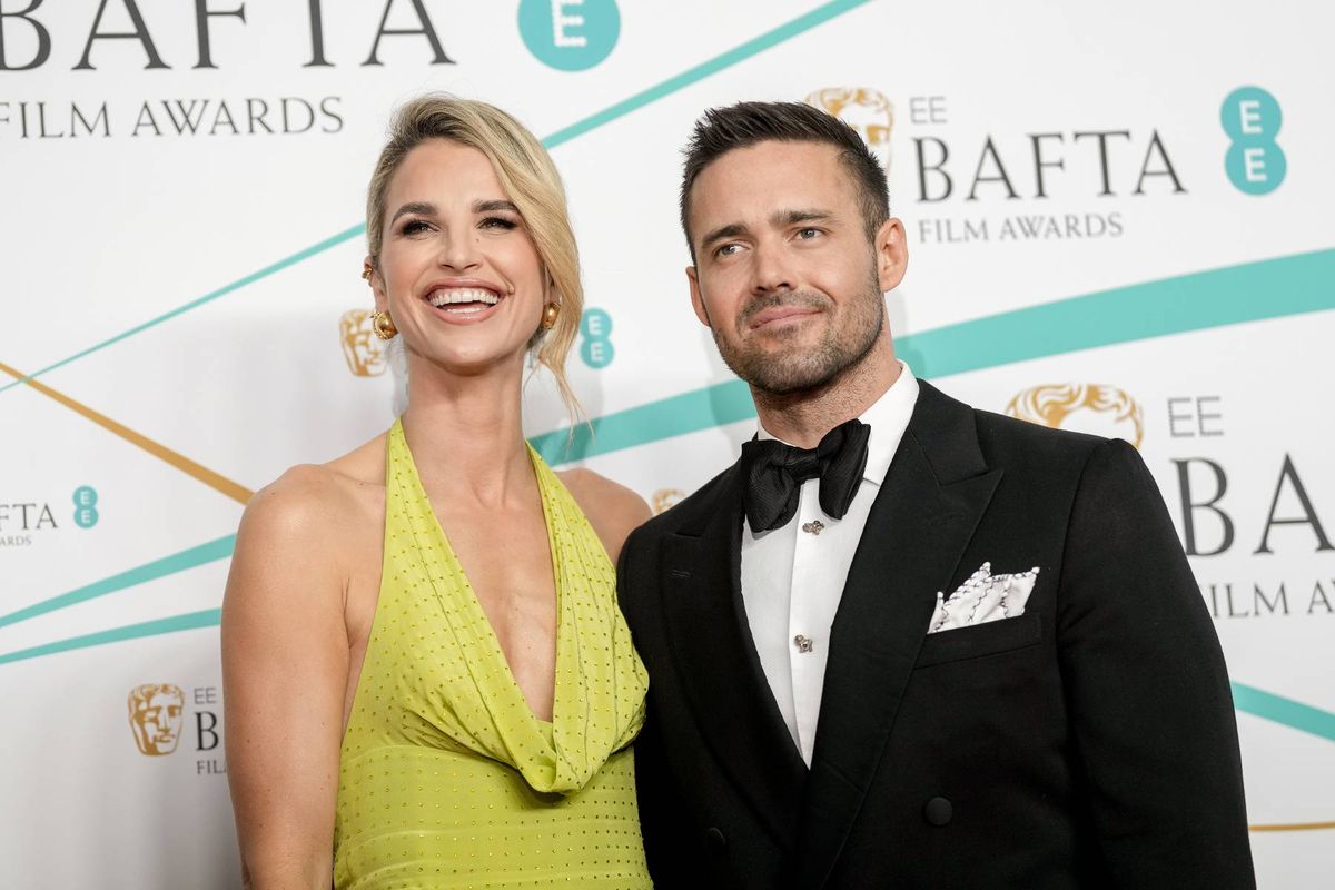 A formal event featuring a man and a woman standing together, likely at the BAFTA Film Awards. The woman is wearing a vibrant yellow dress, and the man is dressed in a black tuxedo with a bow tie. Behind them is a backdrop with the BAFTA logo and event branding.