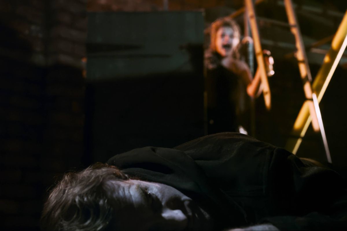 An individual lying on the ground with another individual standing behind them, both appearing to be in a dimly lit area with a yellow light source in the background.