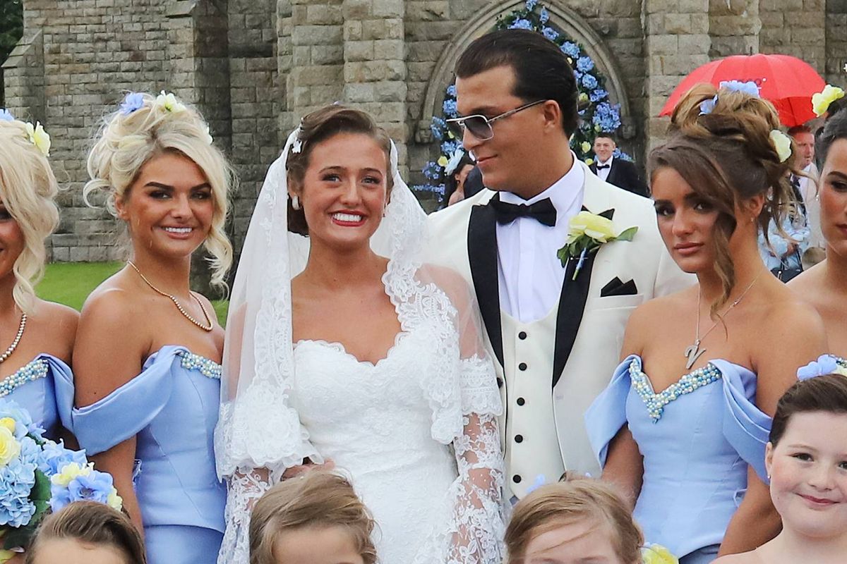 A group of young children, dressed in matching blue dresses, are standing together, possibly in a celebratory setting. Some of them are smiling and looking forward, while others are gazing in different directions. In the background, a woman in a white gown, likely the bride, is surrounded by other individuals, possibly bridesmaids, also dressed in blue.