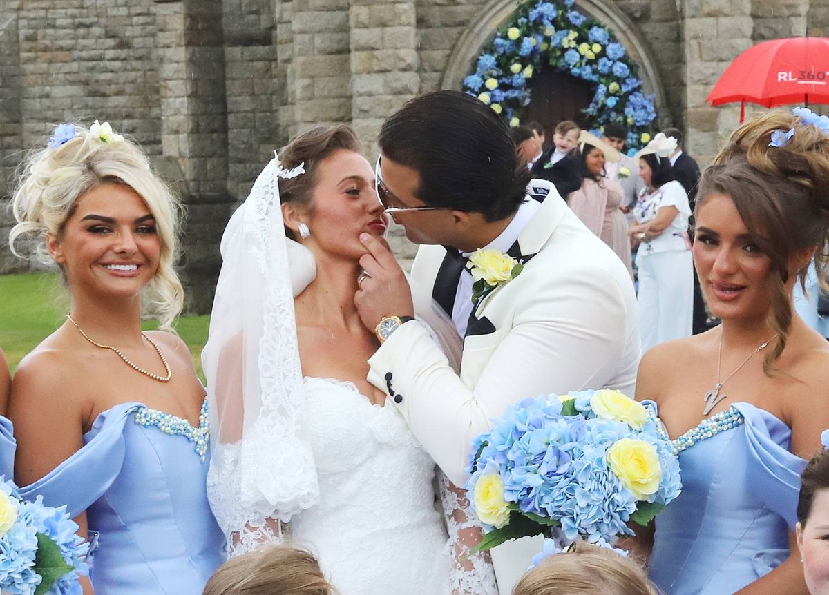 A newlywed couple, the groom in a white suit and the bride in a white gown, shares a tender moment as they are surrounded by bridesmaids wearing blue dresses and holding bouquets. The scene takes place outdoors with a stone building in the background.