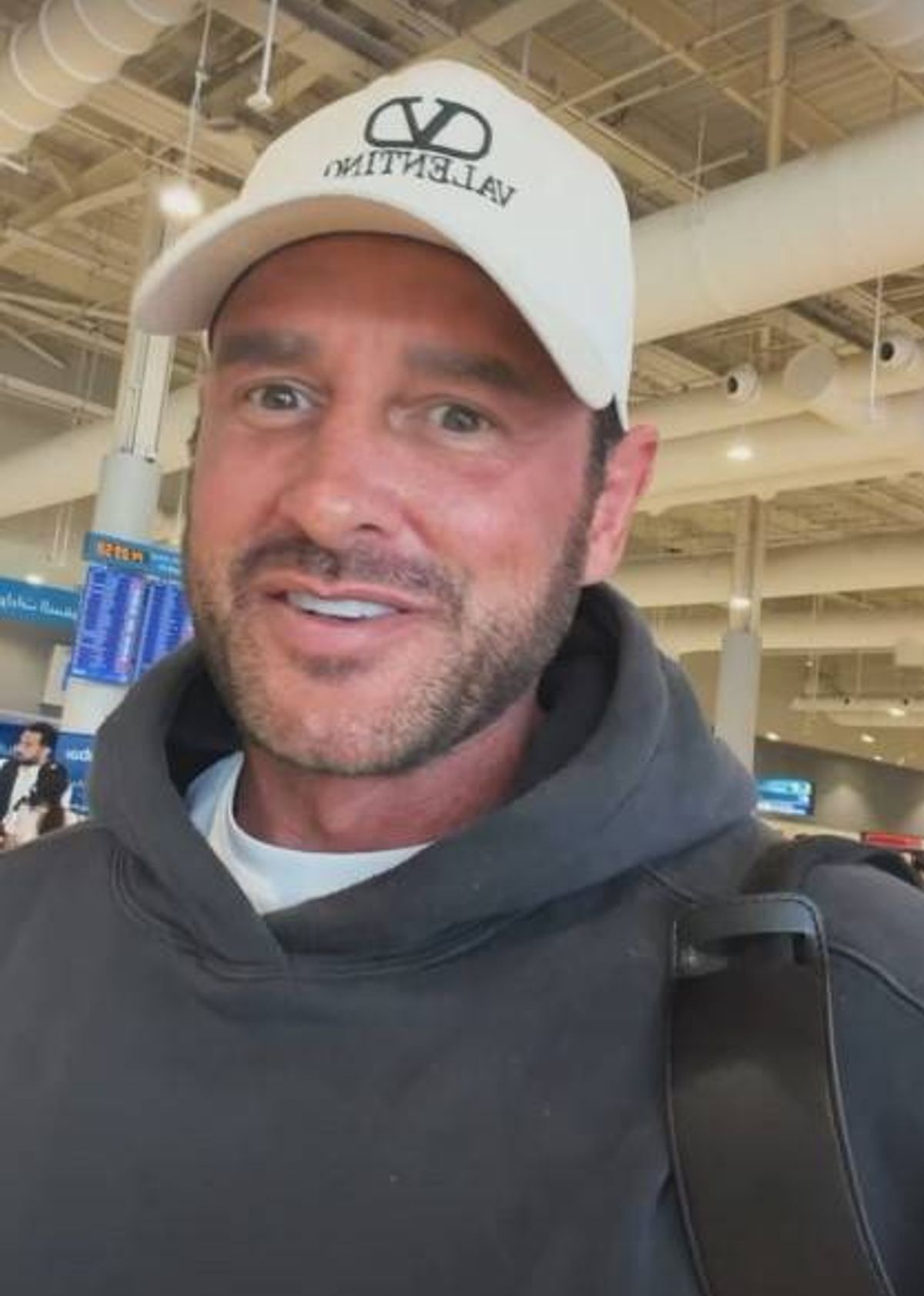 An individual with a beard and mustache, dressed in a white cap and dark hoodie, is captured in a close-up shot at a modern indoor facility, possibly an airport, with an architecturally designed ceiling adorned with lighting fixtures and a metallic structure.