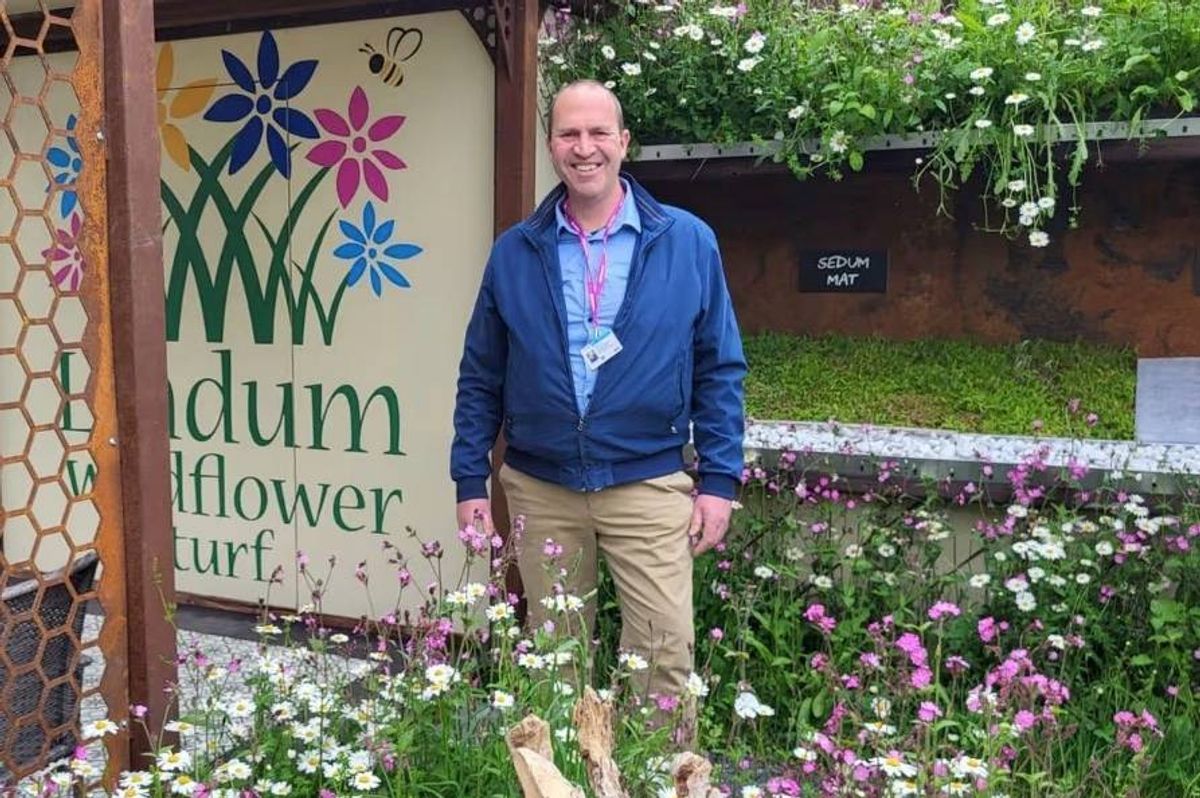 An individual dressed in a blue jacket and khaki pants stands in front of a sign adorned with floral designs, within a vibrant garden setting. The sign, partially obscured by the individual, features the words "Plum flower art" along with decorative elements. The garden is lush with various blooming flowers, and a brick wall with greenery is visible in the background.