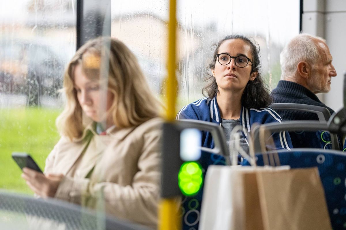 An image depicts three individuals seated on a bus, with one person engrossed in their mobile phone, another person gazing into the distance, and a third person looking downward. The bus interior features a green traffic light, a metal pole, and a window that reveals a grassy area and parked vehicles.