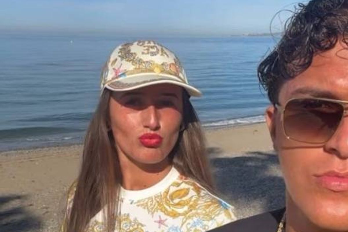Two individuals are standing on a sandy beach near a body of water. The person on the left, wearing a hat and red lipstick, is looking directly at the camera. The person on the right, wearing sunglasses and a necklace, is gazing to the side. The sun casts shadows on the sand behind them.