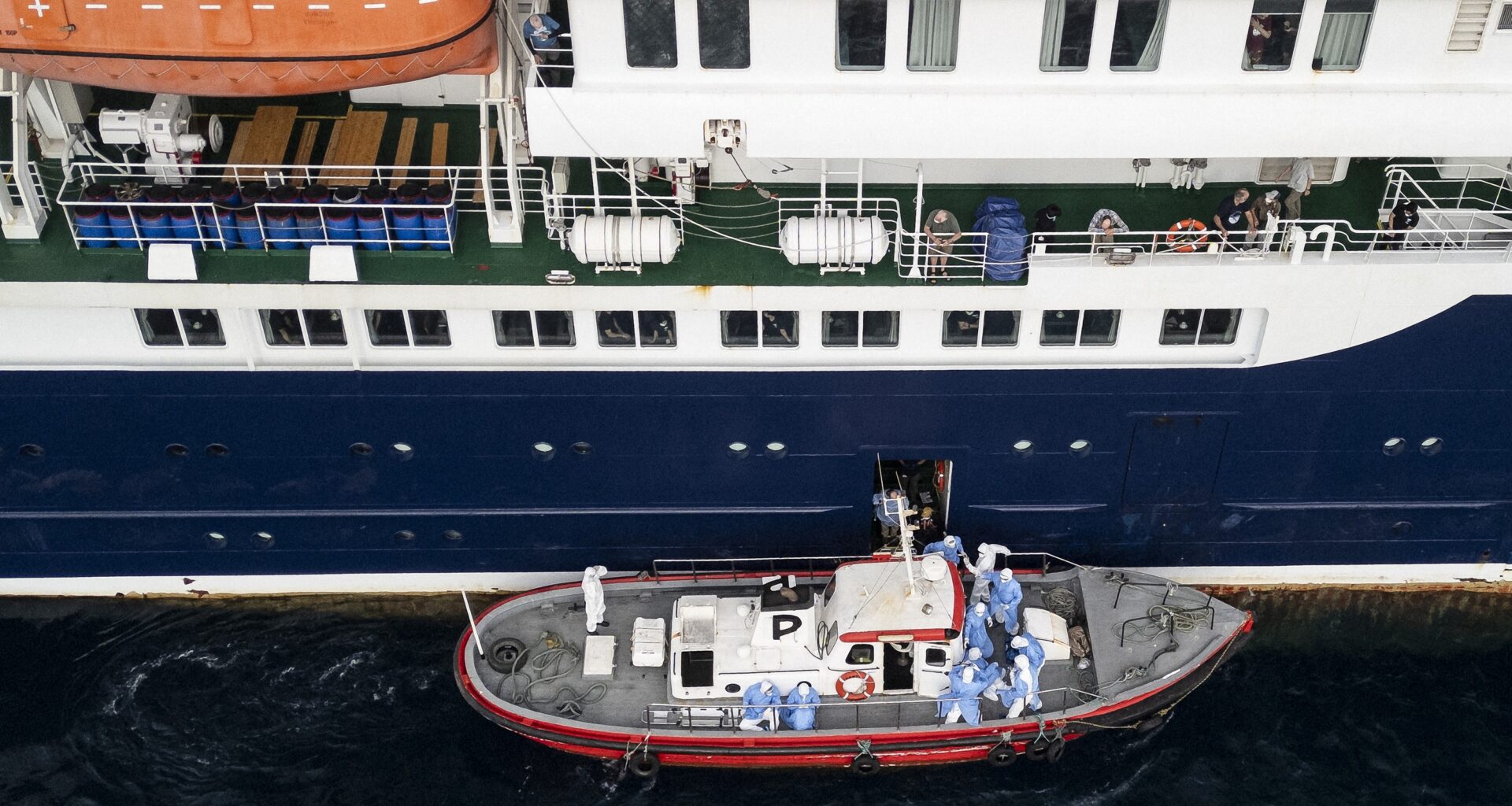 People in blue hazmat suits are on a small boat running alongside a cruise ship.