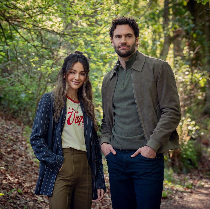 two individuals stand in a wooded area surrounded by greenery two individuals stand in a wooded area surrounded by greenery