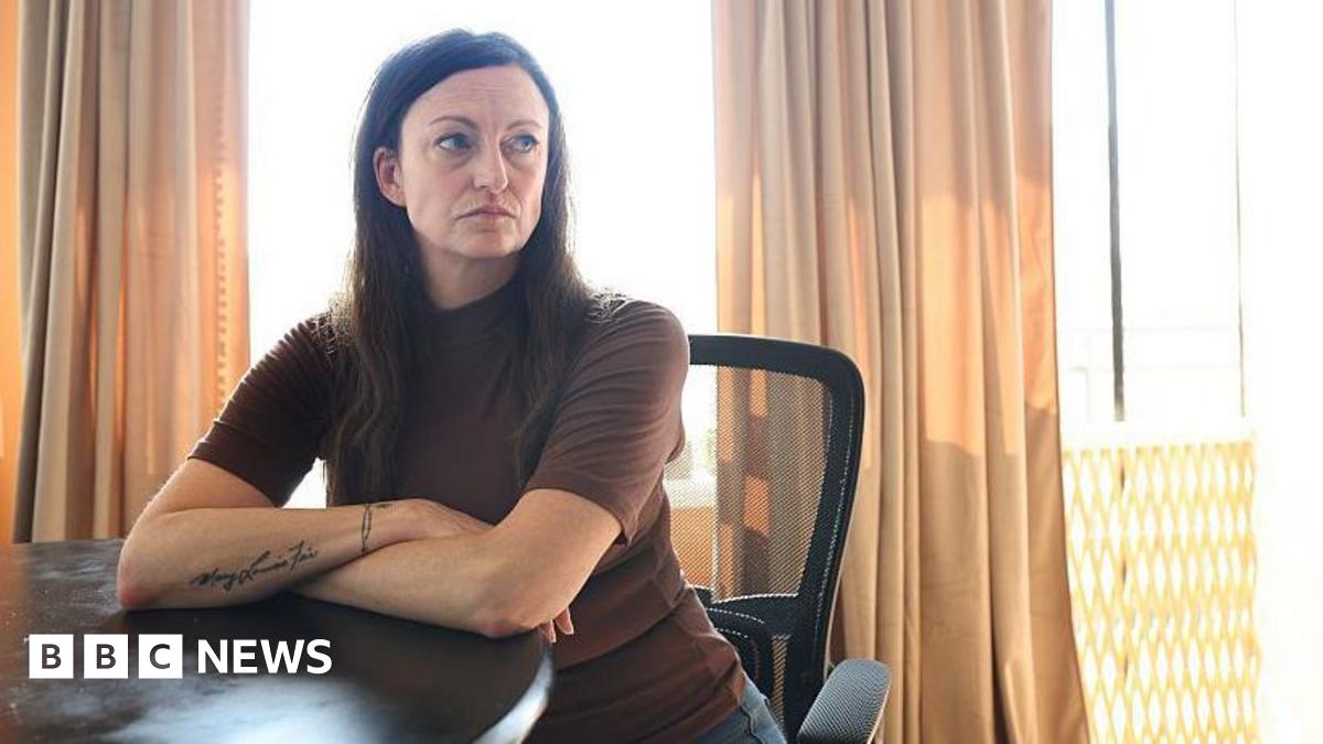 Claire Brosseau is photographed at her home in Toronto, Canada. She has dark brown hair and blue eyes. She is seated at a table with her arms crossed. She is wearing a brown short-sleeved turtle neck.