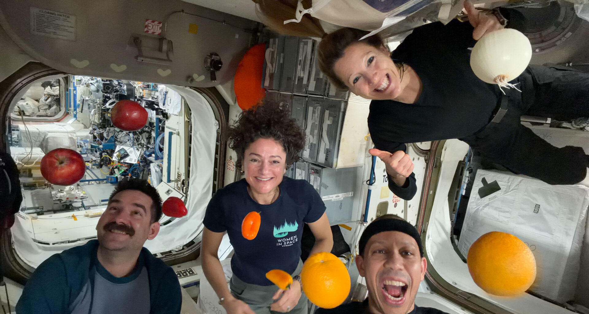 NASA astronauts Jack Hathaway (bottom left), Jessica Meir (middle left), and Chris Williams (bottom right), and ESA (European Space Agency) astronaut Sophie Adenot (top right) pose for a group photo aboard the International Space Station, as fruits and vegetables float around them.