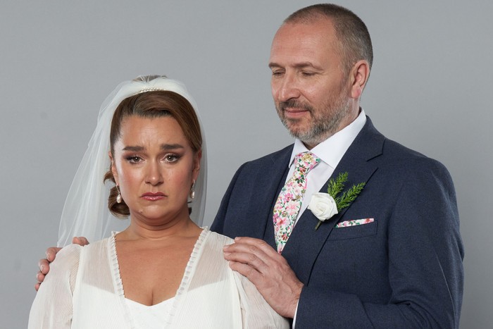 563011,TITLE:Eastenders Ross, in a suit, puts his hand on Vicki's shoulder in key art for EastEnders. She is wearing a white wedding dress and veil.
