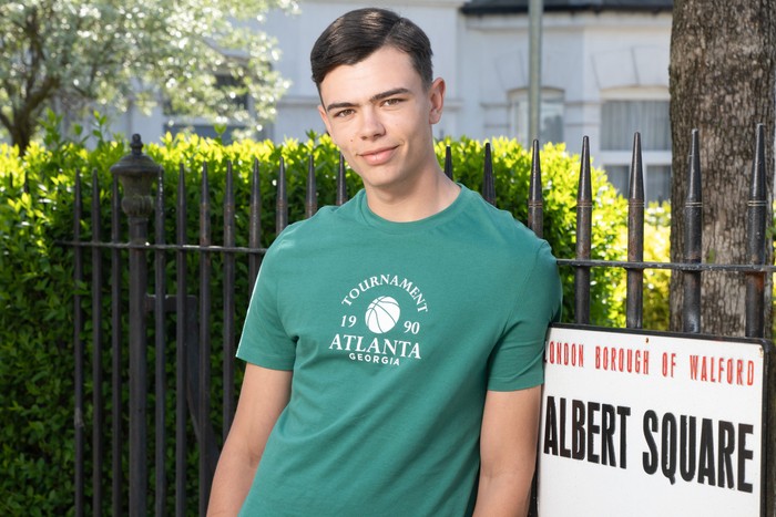 565130,TITLE:Eastenders Will Mitchell, in a green t-shirt, stood in front of the Albert Square sign in EastEnders