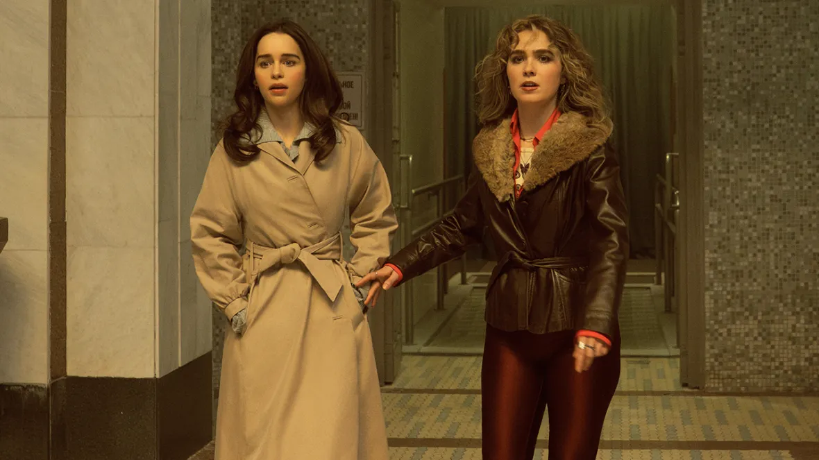 Two women, one in a trench coat and the other in a fur-collared leather jacket, hold hands while looking shocked.