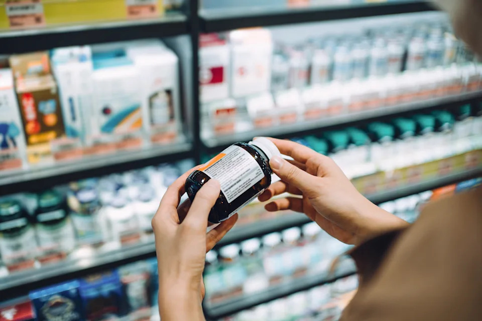 Over the shoulder view of young Asian woman browsing through medical products and reading the label on a bottle of medicine in front of the shelves in a pharmacy