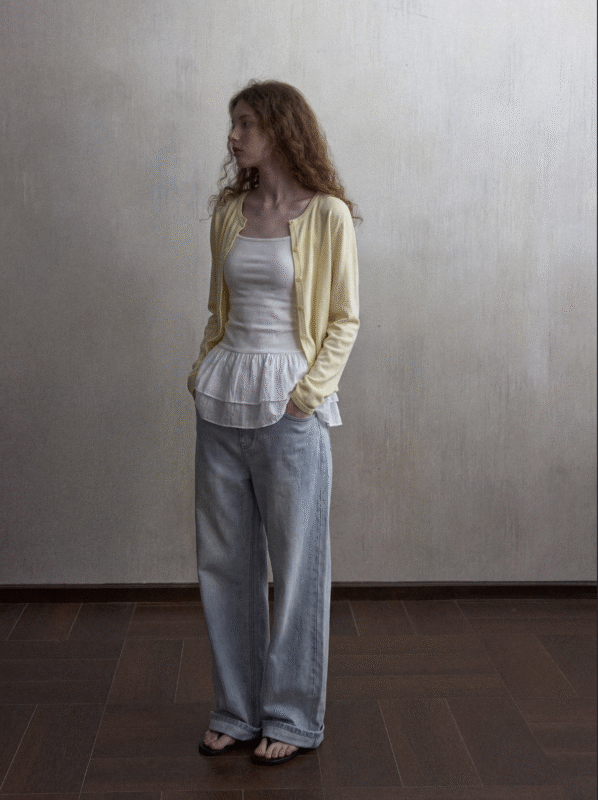 A woman with long curly hair stands indoors, wearing a light yellow cardigan over a white ruffled top, loose light blue jeans, and black sandals, with her hands in her pockets, looking to the side.