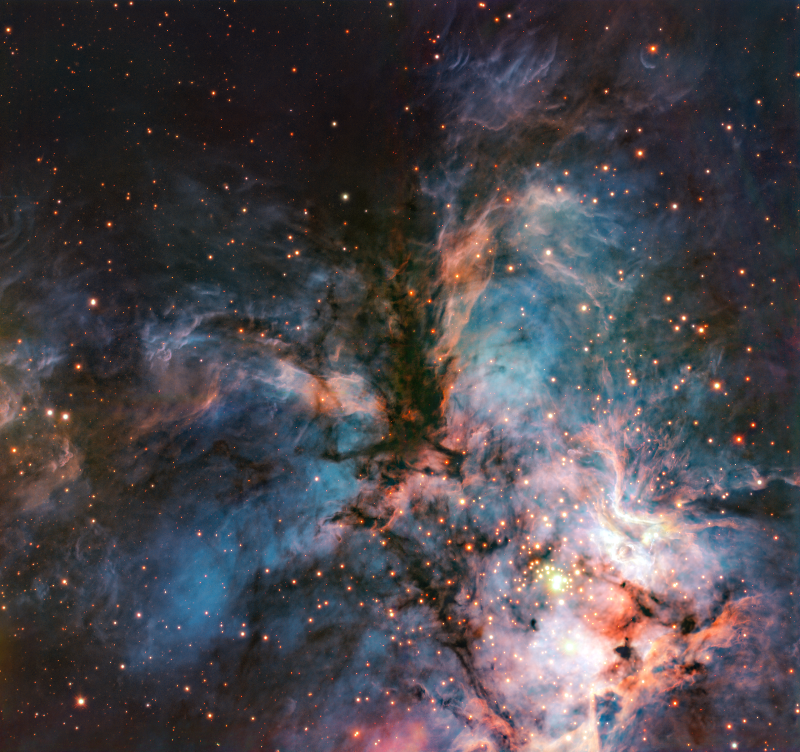 A richly detailed telescope image of a star-forming nebula, filled with swirling clouds of blue, orange, and pink gas and dust. Dark pillars of dense dust cut through the center of the image, surrounded by glowing nebular material and hundreds of orange and yellow stars scattered throughout. A bright cluster of young stars illuminates the lower right portion of the image, casting a warm glow against the surrounding gas clouds.