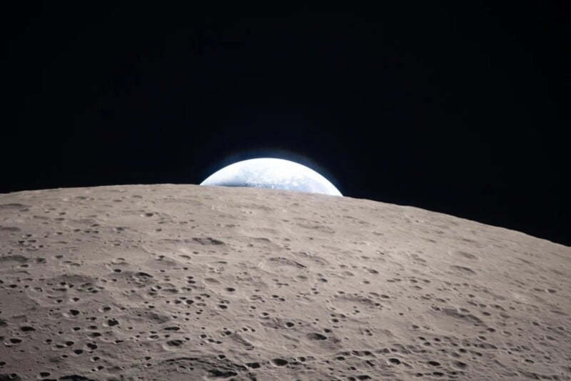 The image shows Earth rising over the cratered gray surface of the Moon, with the bright blue and white Earth appearing on the dark horizon against the blackness of space.