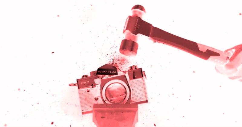 A hammer is about to smash a vintage Praktica MTL 5 camera, with debris and particles flying around. The image has a red tint and high contrast.
