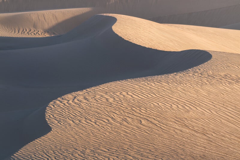 Curving sand dunes cast soft shadows in the sunlight. The surface of the sand is textured with fine, wavy patterns caused by the wind. The light and shadow highlight the smooth contours of the landscape.