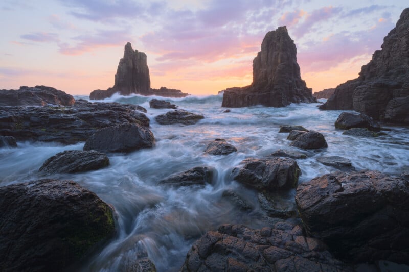 Rocky coastal landscape at sunrise, with waves crashing against dark rocks and tall sea stacks under a colorful sky with pink and orange clouds.