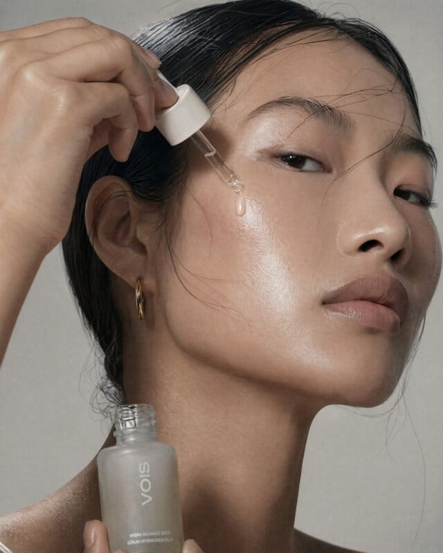 A woman with dewy skin applies serum to her face using a dropper. She holds a bottle labeled "VOIS" and has her hair slicked back, wearing a gold hoop earring and a natural makeup look.