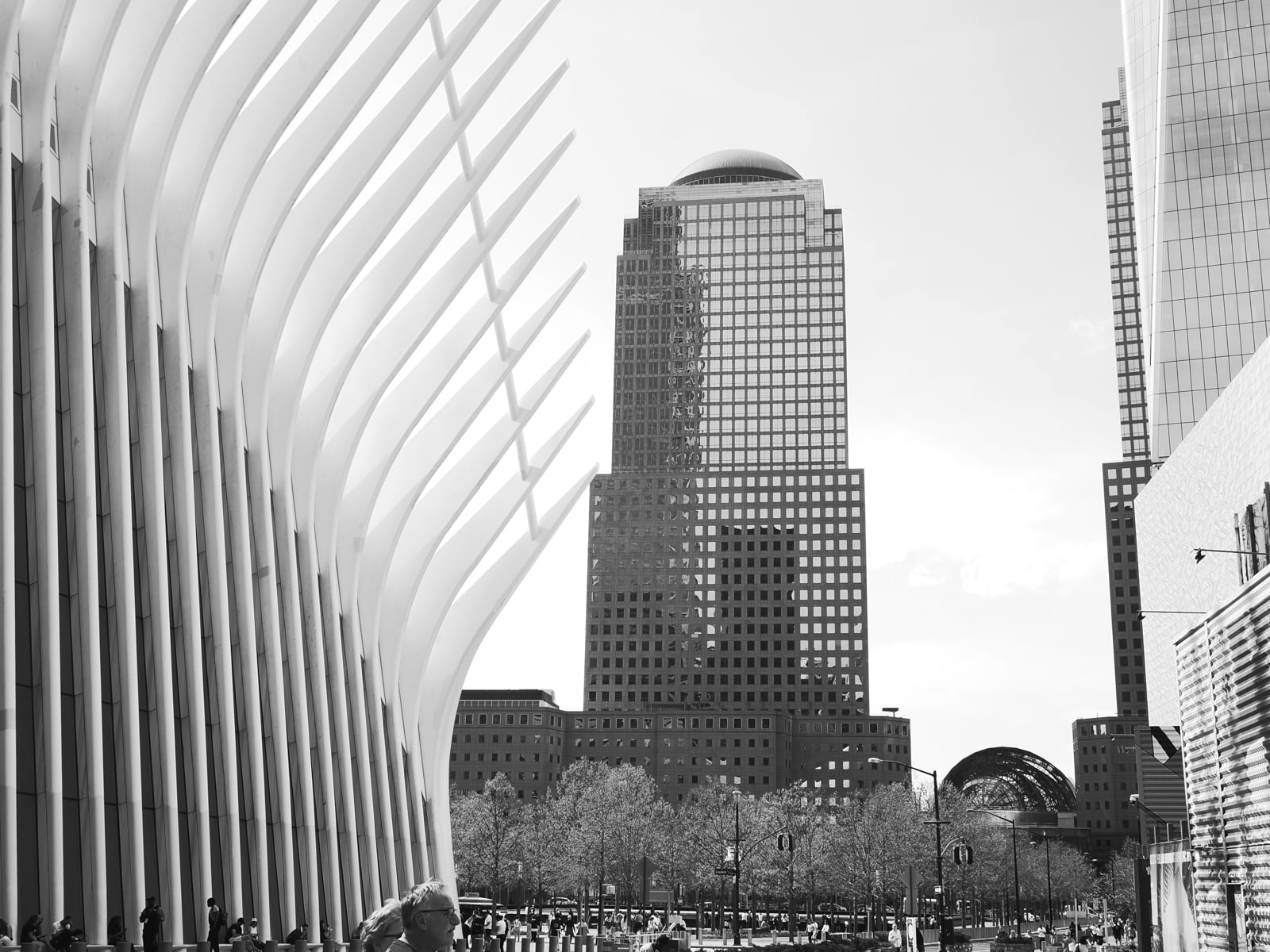 Black and white photo of the Oculus structure at the World Trade Center in New York City, with tall buildings and trees in the background, and people walking in the foreground.