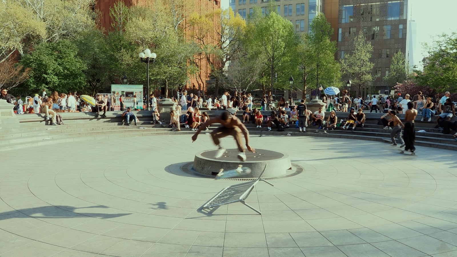 A skateboarder performs a trick over a circular fountain in a busy urban plaza, while a shopping cart lies tipped over nearby. People sit and stand around the square, watching and enjoying the sunny day.