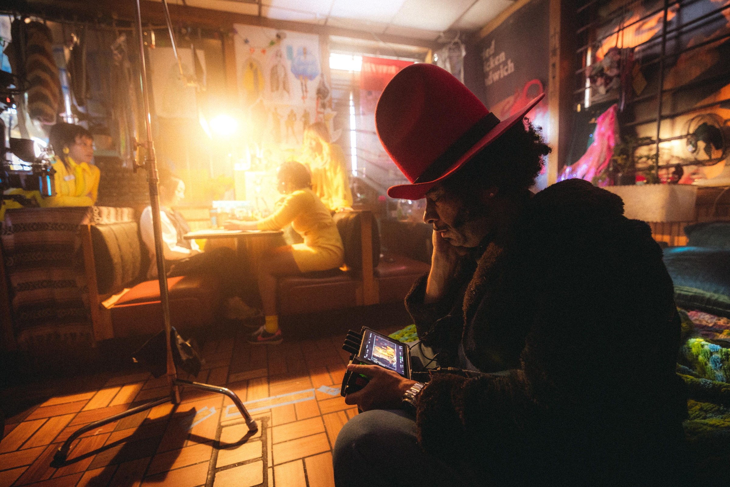 A man in a fur coat and a large red hat sitting on a film set that looks like a diner. The man is holding an electronic device.