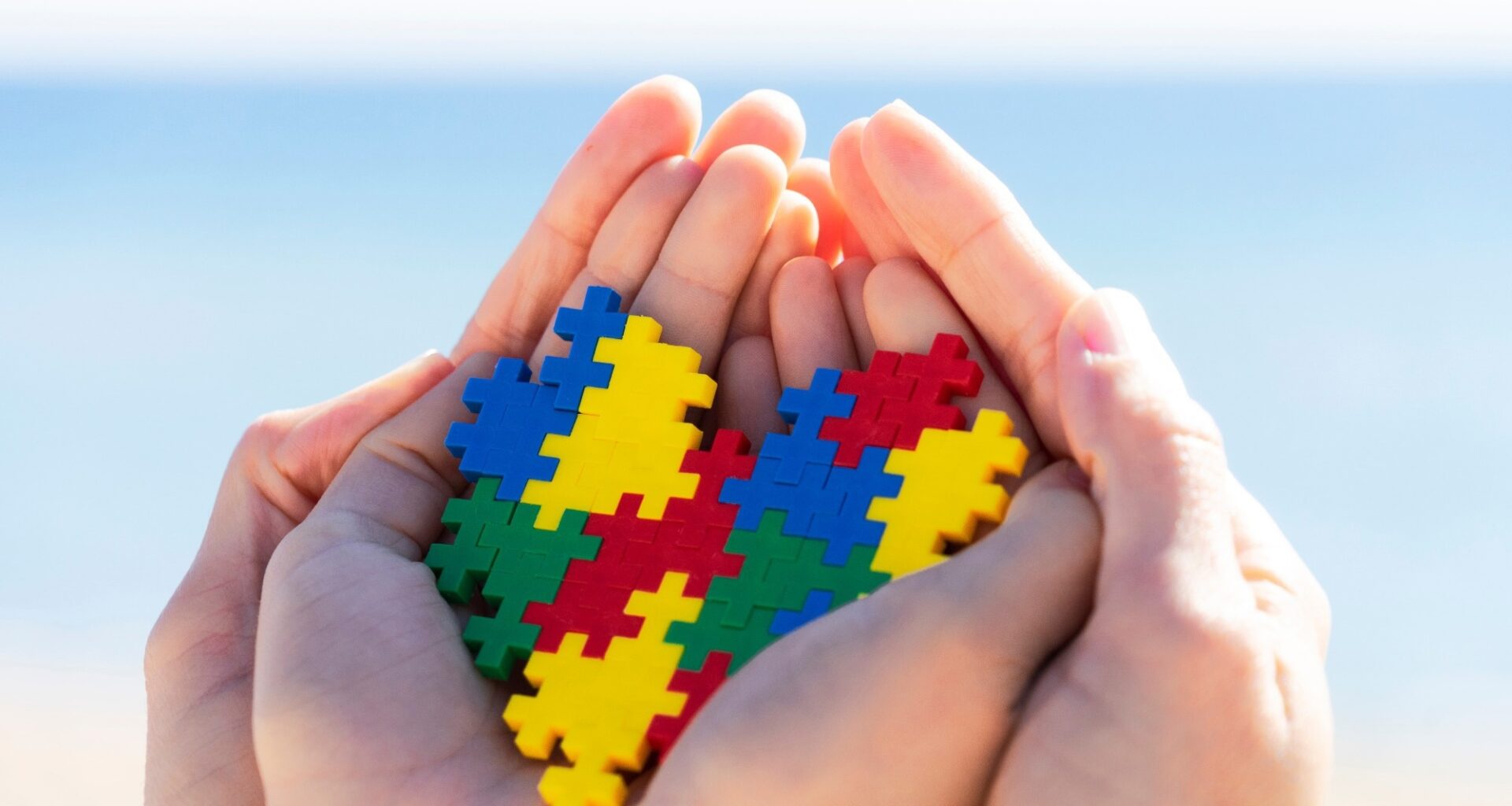 Child and adult hands holding colorful heart with blue background outdoors.