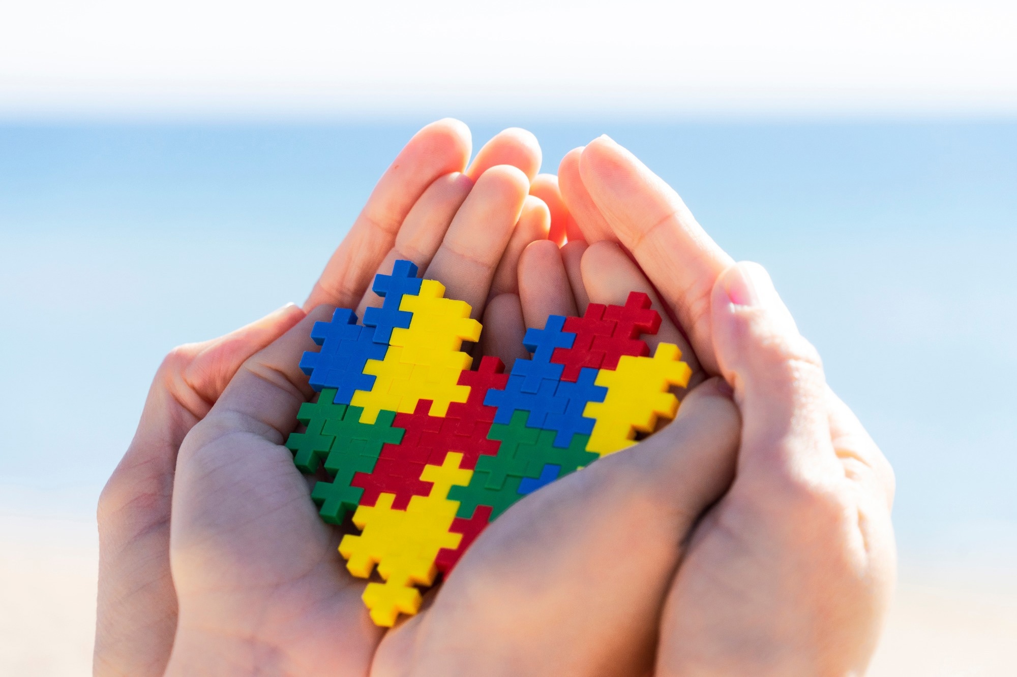 Child and adult hands holding colorful heart with blue background outdoors.