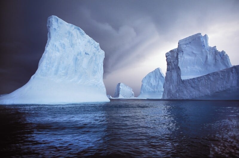 Large icebergs float in dark blue water beneath a dramatic, cloudy sky. The towering ice formations are white and blue, contrasting sharply with the water and moody atmosphere.