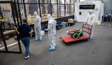 Image shows three people in white protective suits inside a room with another person wearing a blue shirt and blue jeans at NASA's Kennedy Space Center in Florida in 2022. Around them is a red cart with a silver structure and green looking wheels. Photo credit: NASA/Kim Shiflett