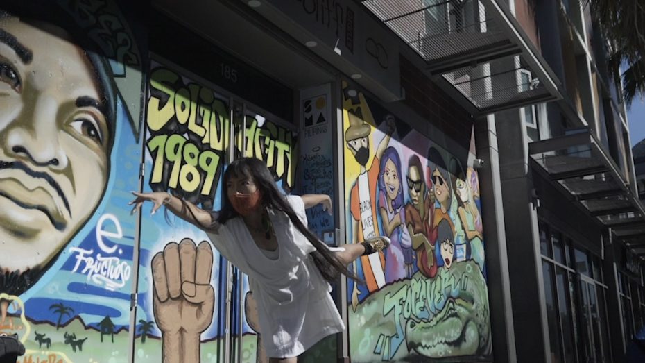 A person poses on one leg in front of a colorful mural featuring portraits, a raised fist, and text reading "SOLU WIDGETS 1989.