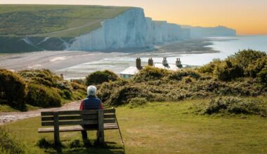Rear view image depicting a senior man in his 70s sitting on a bench leading down to the iconic Seven Sisters cliffs on the coastline of East Sussex, UK. The man is wearing casual clothing - blue denim jeans, a red checked shirt, navy blue gilet. The man is having a rest from hiking and his hiking pole is leaning up against the bench.
