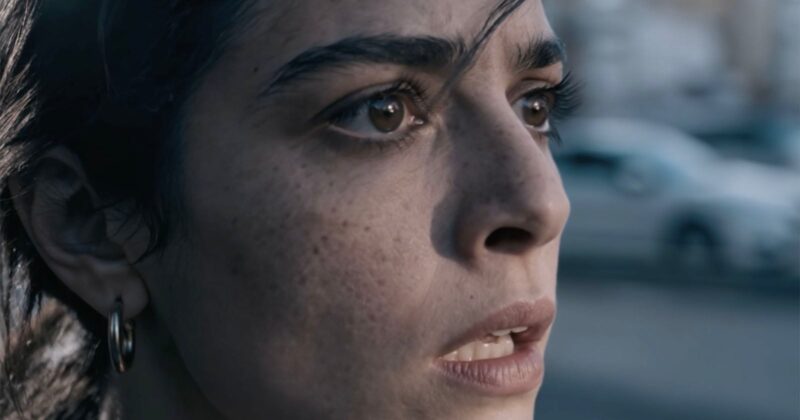 A close-up of a woman with dark hair and brown eyes, looking ahead with a worried or anxious expression. Her mouth is slightly open, and the background appears blurred, showing an outdoor scene.