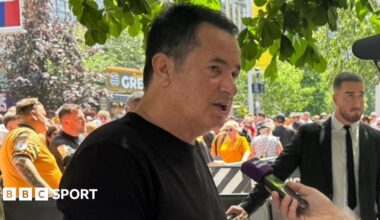 Hull City owner Acun Ilicali being interviewed outside Wembley Stadium, wearing a black t-shirt. Crowds on Olympic Way can be seen behind him.