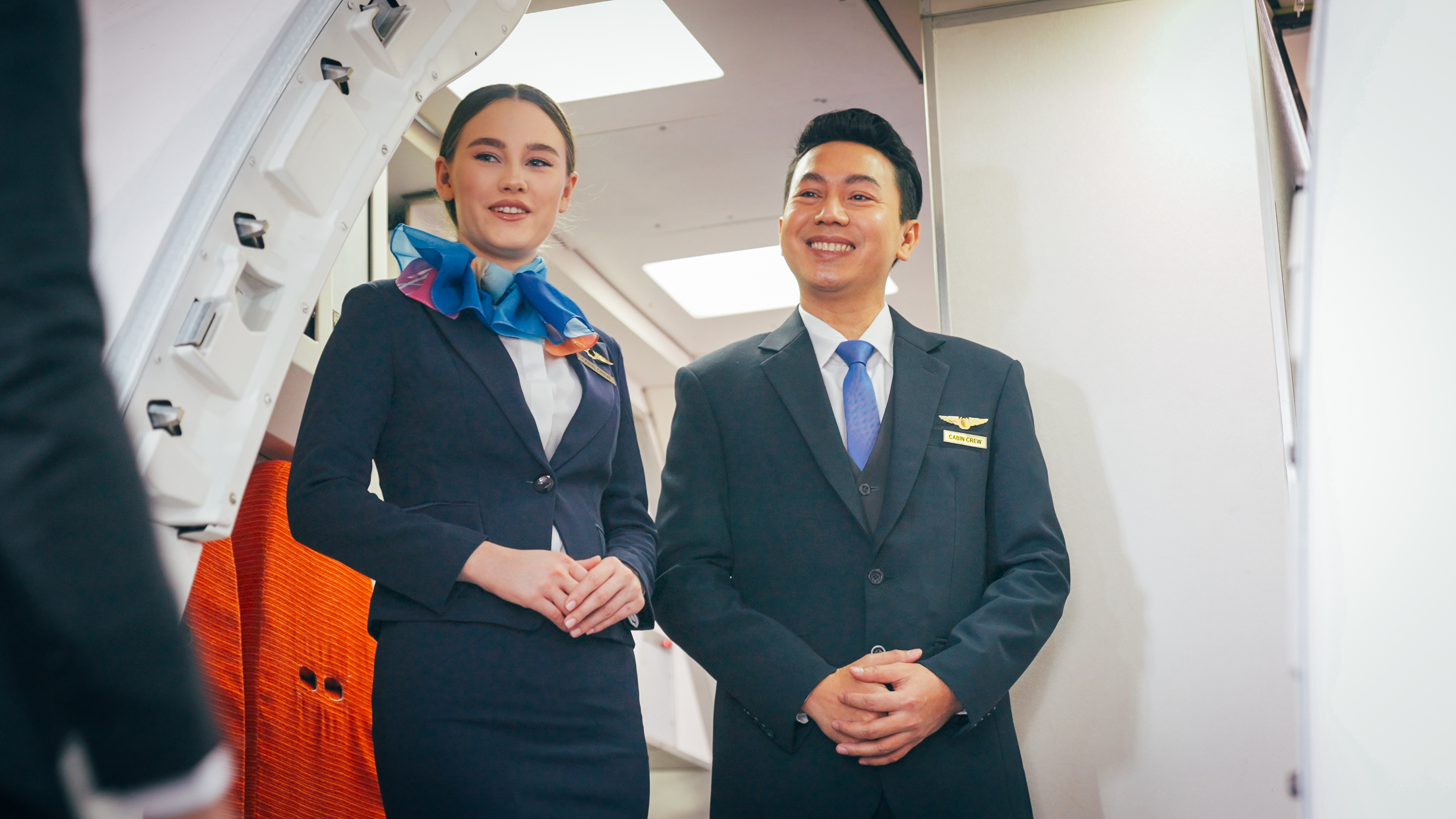 Two smiling cabin crew members, a woman and a man, greet passengers at the airplane entrance.