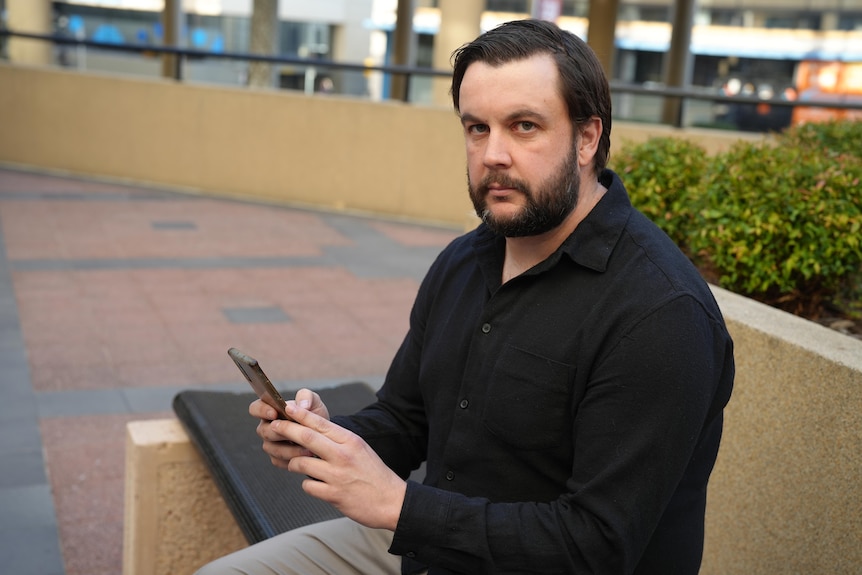 Man in black shirt sits on bench with serious face