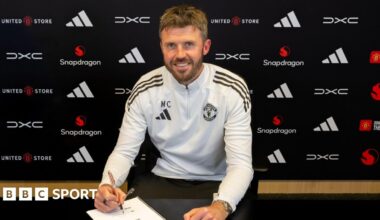Michael Carrick sat down in Manchester United training kit, posing holding a pen over a contract document