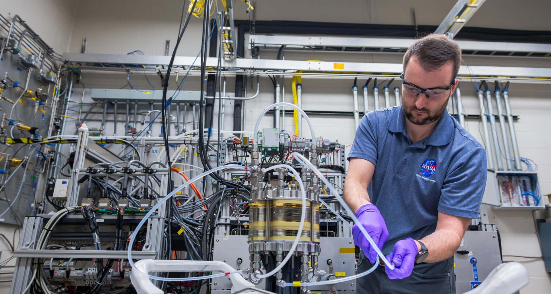 A man wearing a blue NASA polo, purple gloves, and safety glasses adjusts a small tube connected to a silver and yellow cylinder inside the Fuel Cell Testing Laboratory. Behind him are numerous other tubes, wires, and sensors connected to a large system and the lab’s wall.