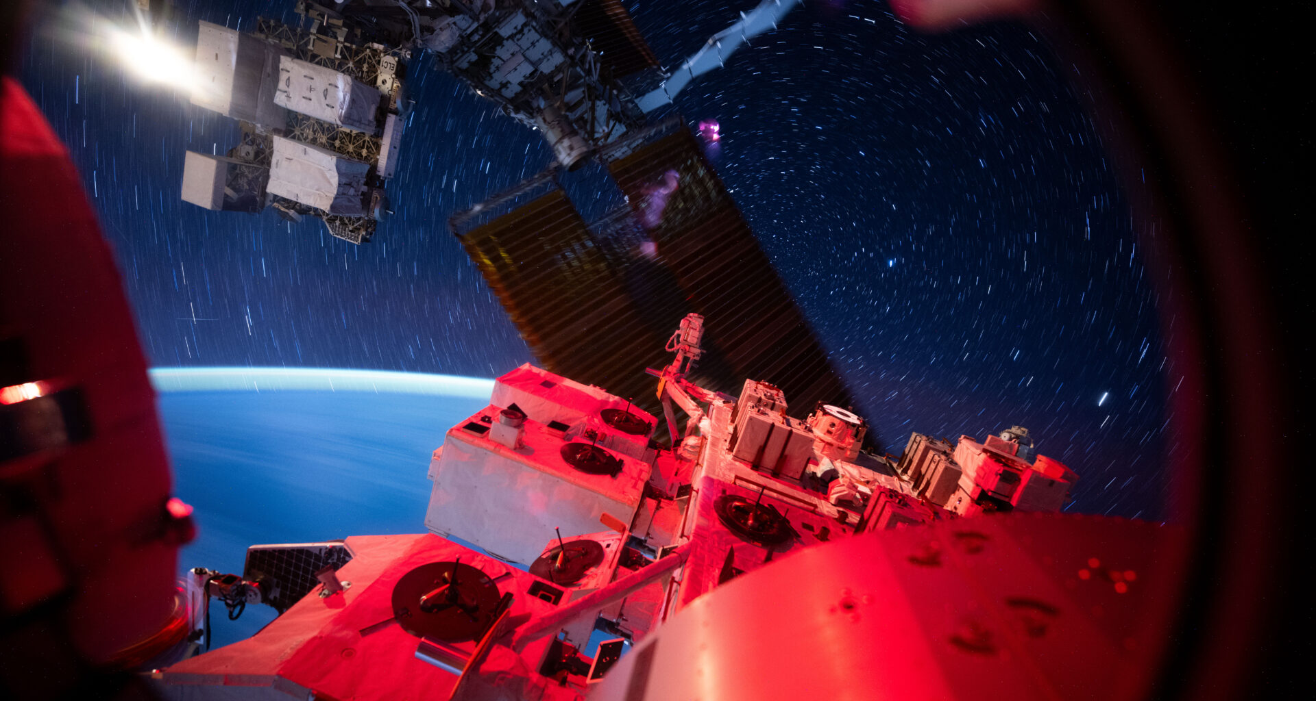 View from the International Space Station showing its illuminated exterior hardware in red light against a backdrop of deep space. Star trails form circular streaks across the sky due to the station’s motion, while Earth’s blue curvature glows softly at the horizon.