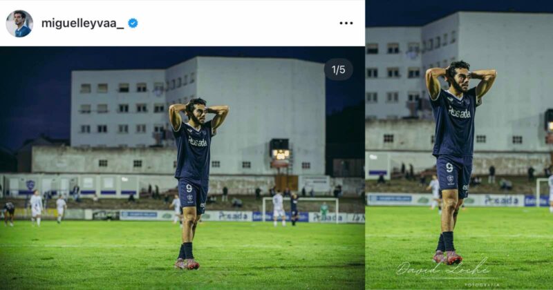 A soccer player wearing a navy blue jersey with the number 9 stands on a field with his hands behind his head during an evening match. Buildings and other players are visible in the background.