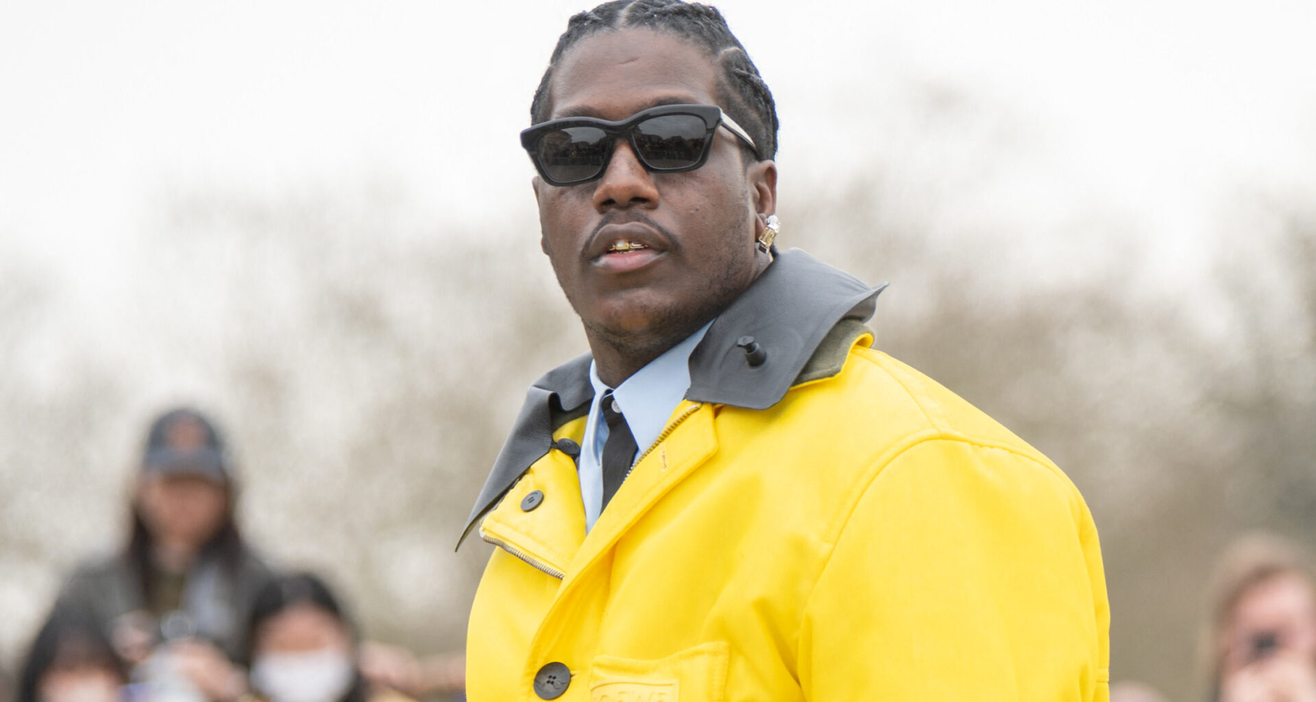 Lil Yachty in a yellow jacket and dark pants holds a blue bag, with a crowd in the background.