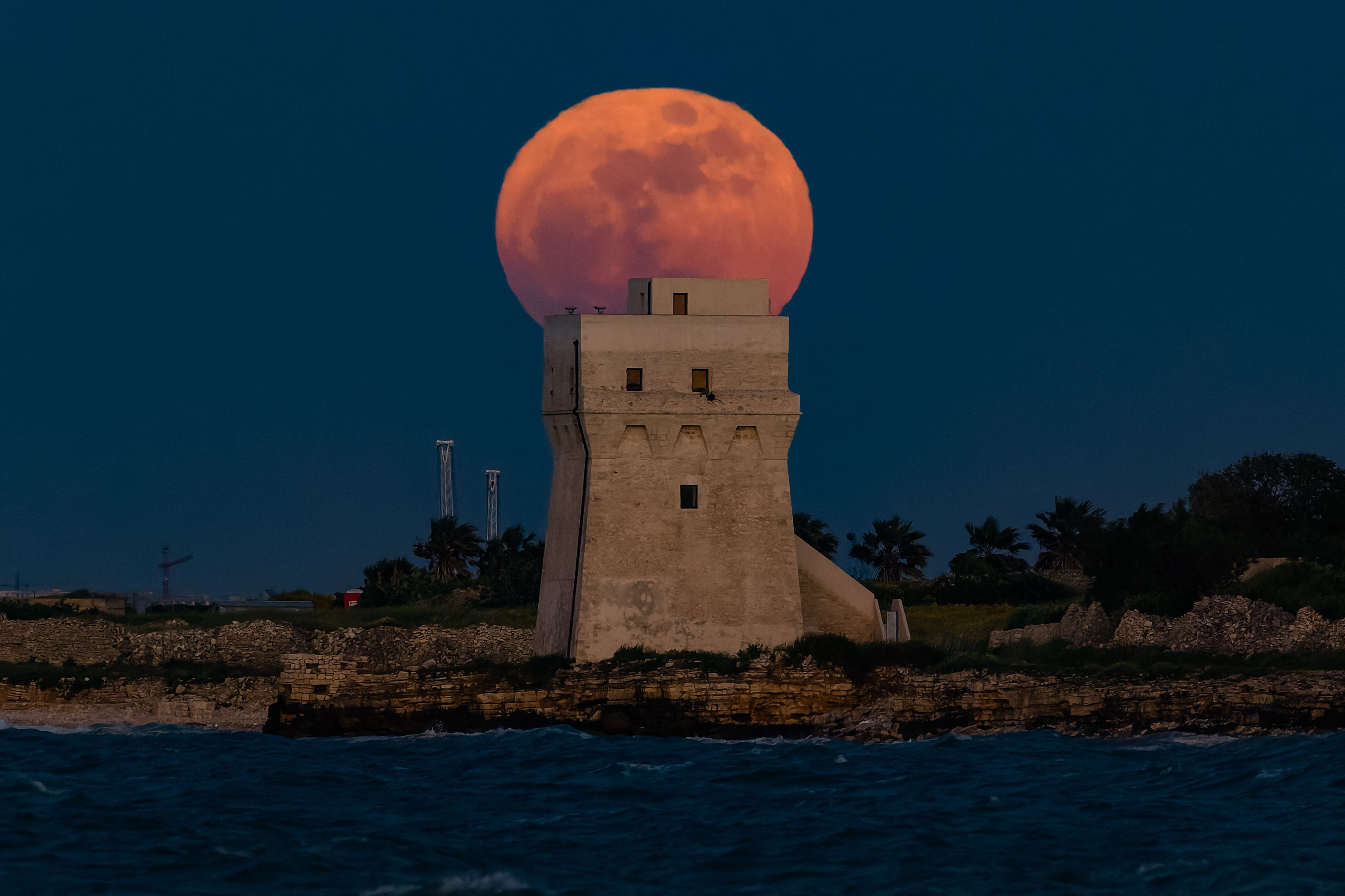 A red full moon is photographed rising behind a stone coastal tower at night.