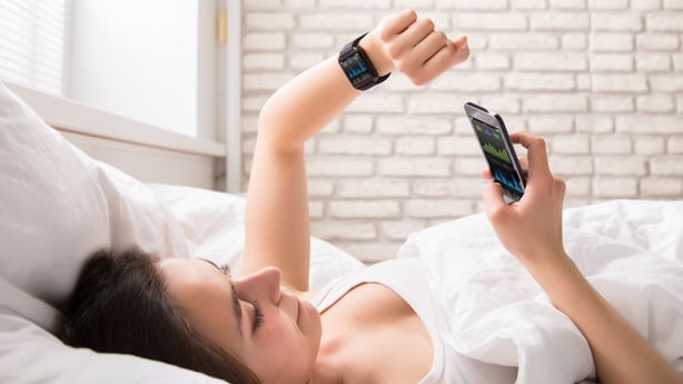 Close-up Of A Young Woman Lying On Bed Synchronizing Smart Watch With Cell Phone
