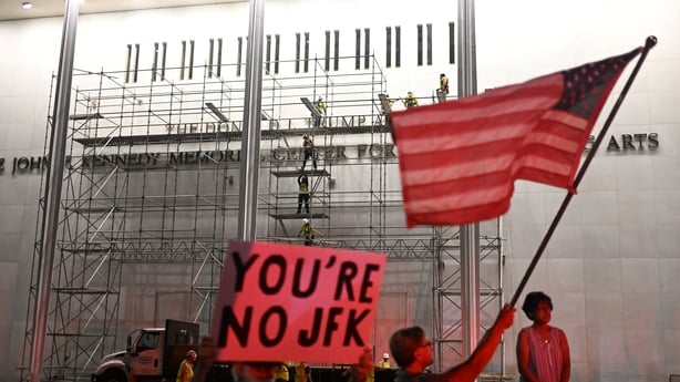 Protesters wave a US flag and hold a sign reading "you're no JFK"