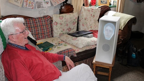 Irish storyteller Brian Caball sits in his living room and speaks with a robot - a white rectangular box with a 3D face