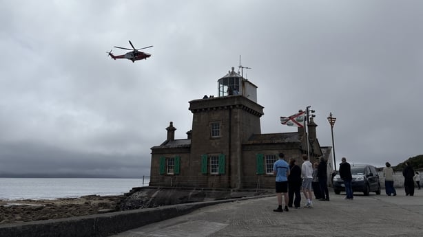 People look up as a helicopter flies above a lighthouse