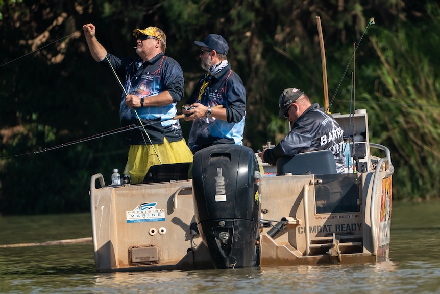 Three men on a small fishing boat, on green river, green trees behind them, two standing, holding lines and one sitting