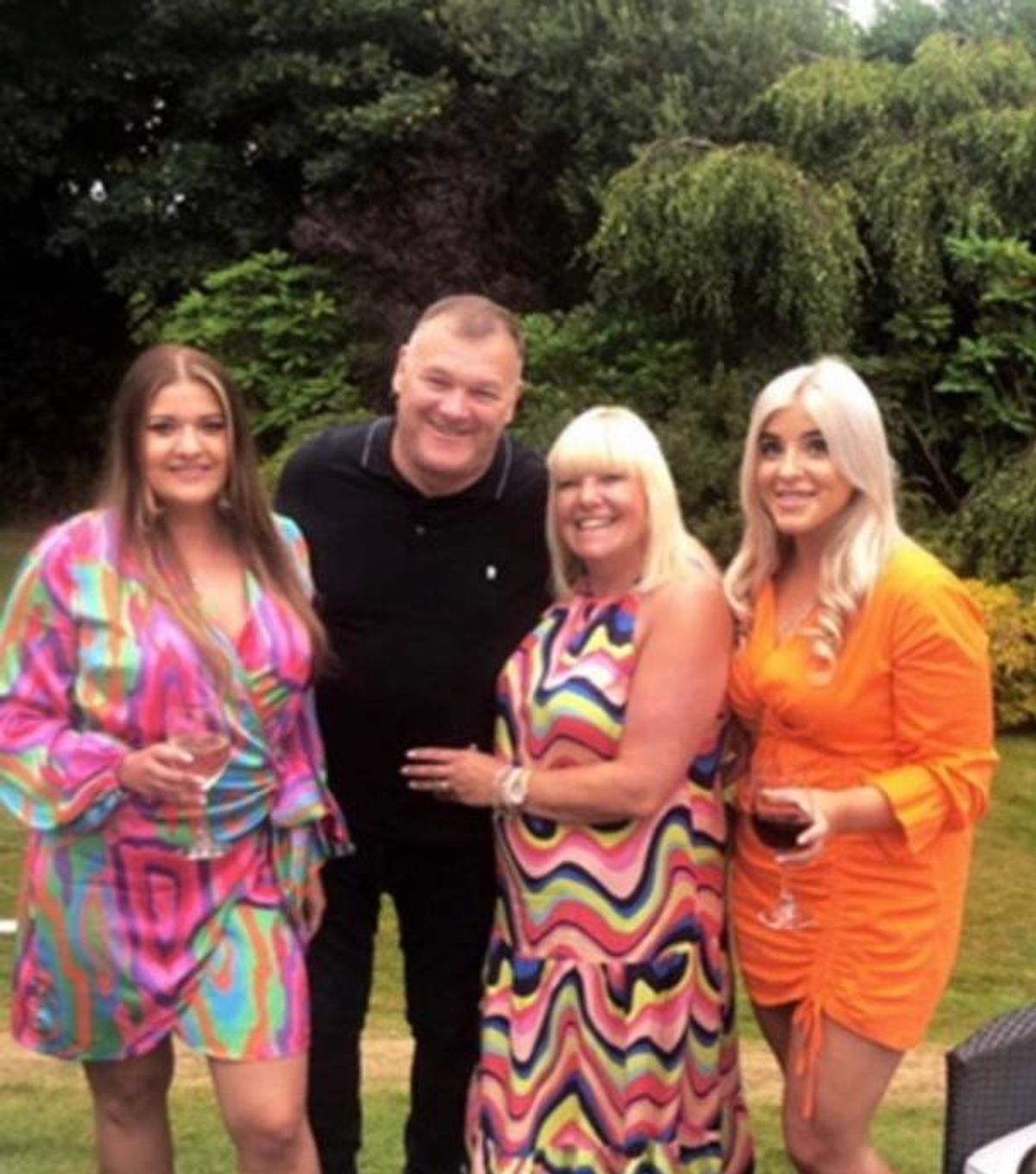 Four individuals are standing together in an outdoor setting, smiling and posing for a photograph. They are dressed in vibrant and colorful outfits, with one individual in the center wearing a black outfit. The background features a lush green lawn and some trees.